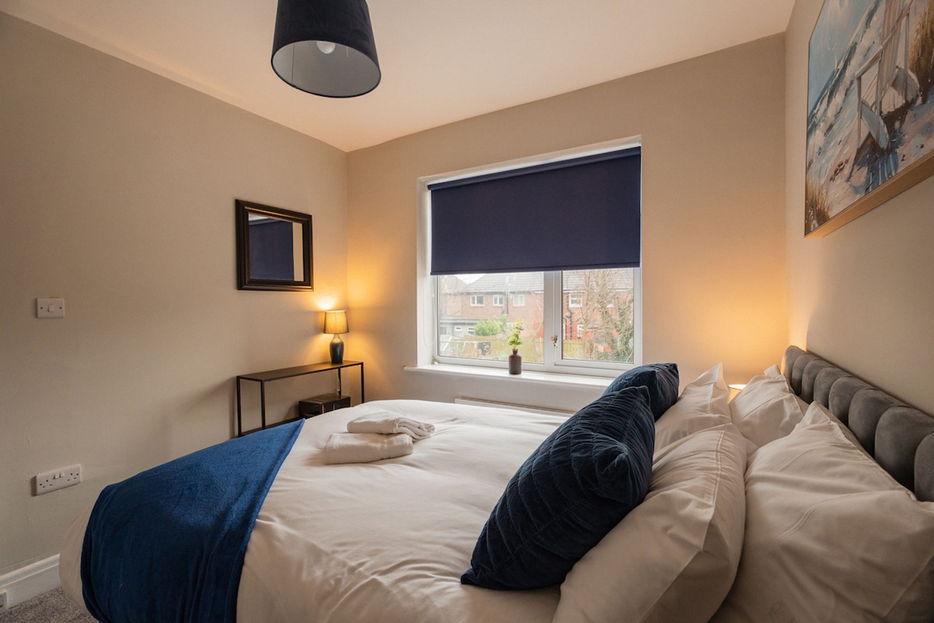 A hotel room with a bed, a window with a view, a painting on the wall, and a lamp on a bedside table.