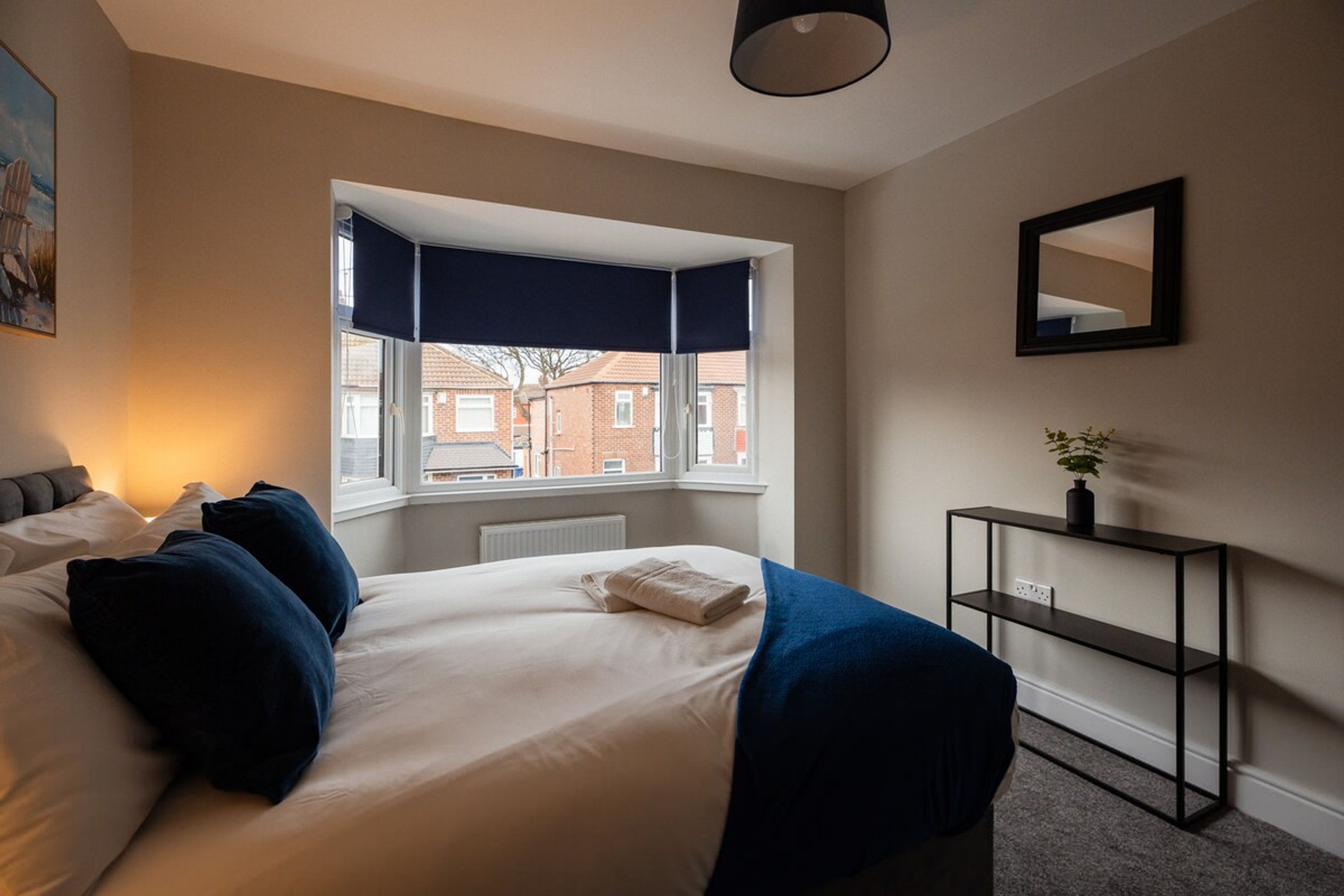 A bedroom with a bed, a window with a view of buildings, a bedside table with a plant, and a mirror on the wall.