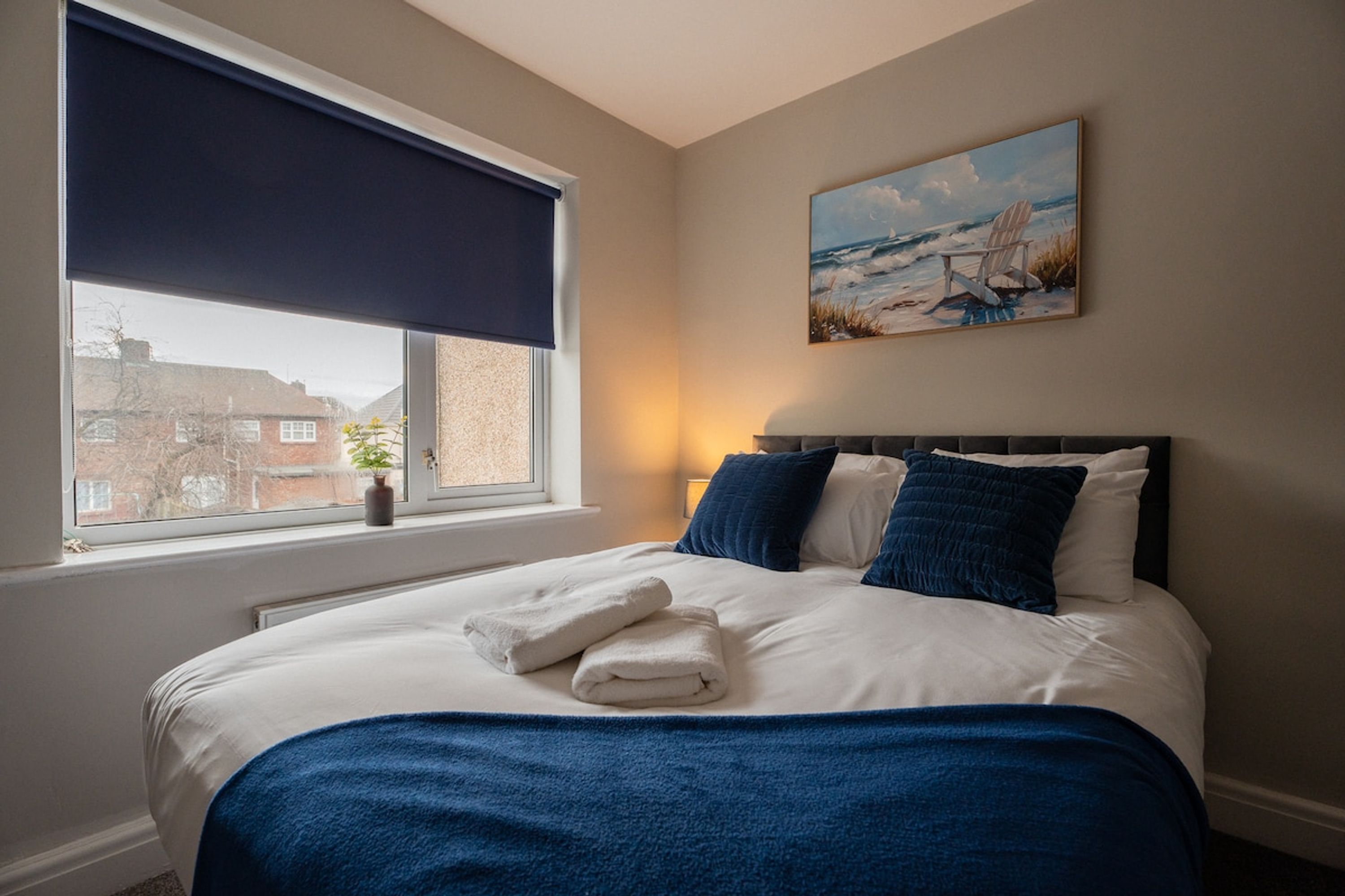 A neatly made bed with white and blue bedding, a window with a dark blind, and a painting of a beach scene on the wall.