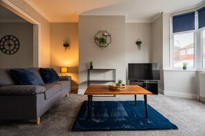 Uma sala de estar com sofá, mesa de centro, televisão e janela com persianas.