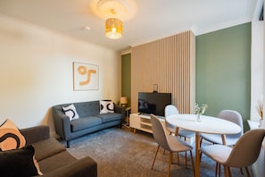A modern living room with a sofa, a dining table with chairs, a flat-screen TV, and a decorative wall panel. - Rockliffe House Horizon Stays (Middlesbrough)