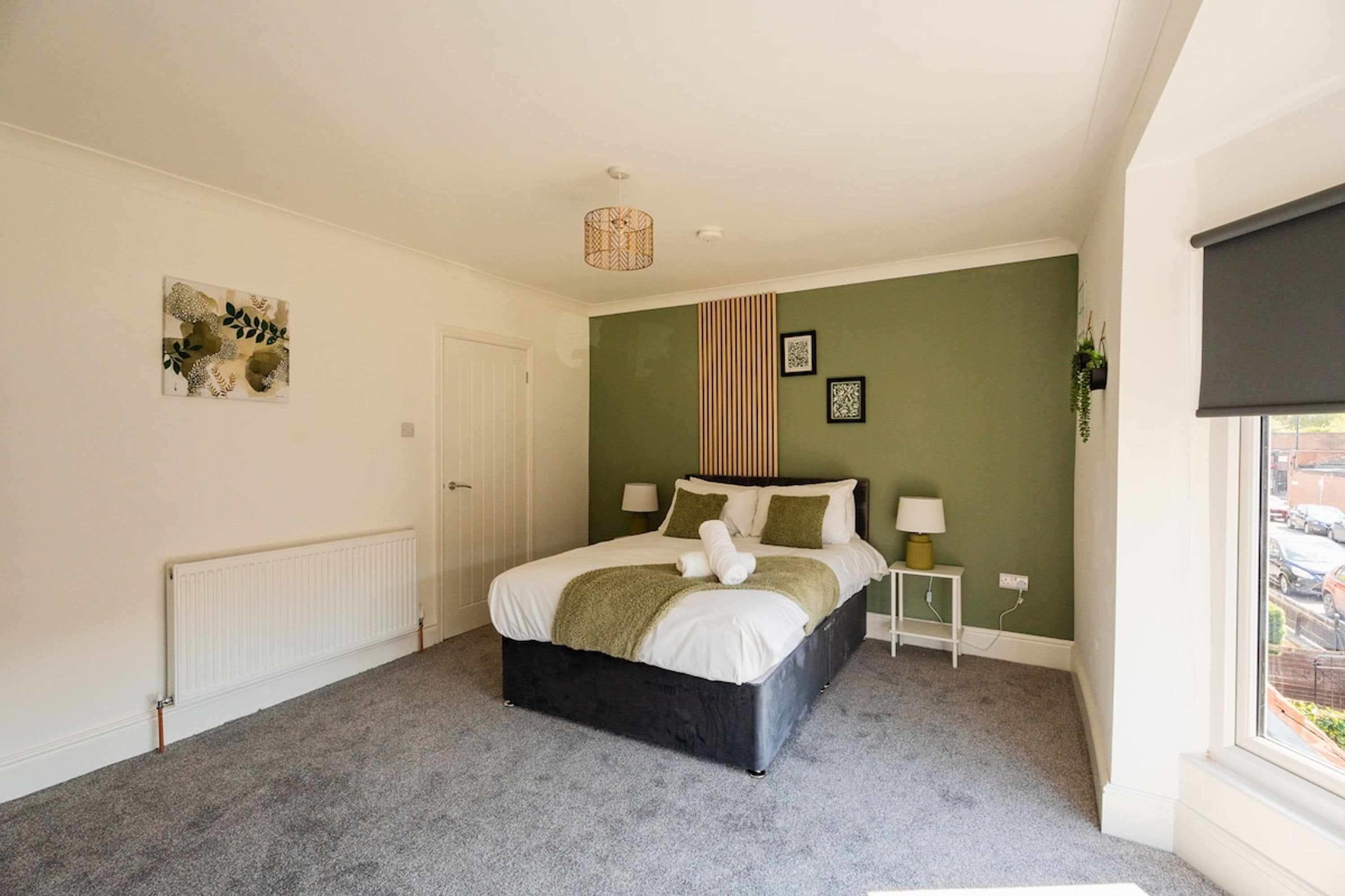A bedroom with a bed, a bedside table, a radiator, a window with a blind, and a wall with a green panel and two framed pictures.