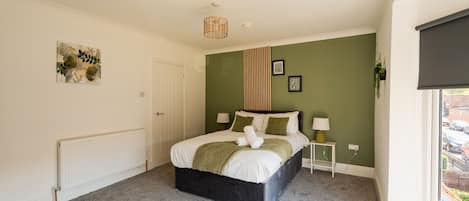 A bedroom with a bed, a bedside table, a radiator, a window with a blind, and a wall with a green panel and two framed pictures.