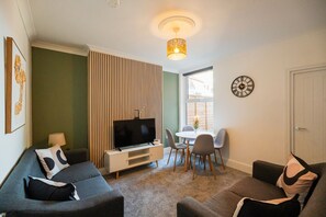 Ein modernes Wohnzimmer mit Flachbildfernseher, einem Essbereich mit kleinem Tisch und Stühlen sowie einer wandmontierten Uhr.
