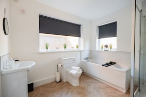 Ein modernes Badezimmer mit Toilette, Waschbecken und Badewanne. Zwei Fenster mit Jalousien und eine kleine Ablage mit Pflanzen.
