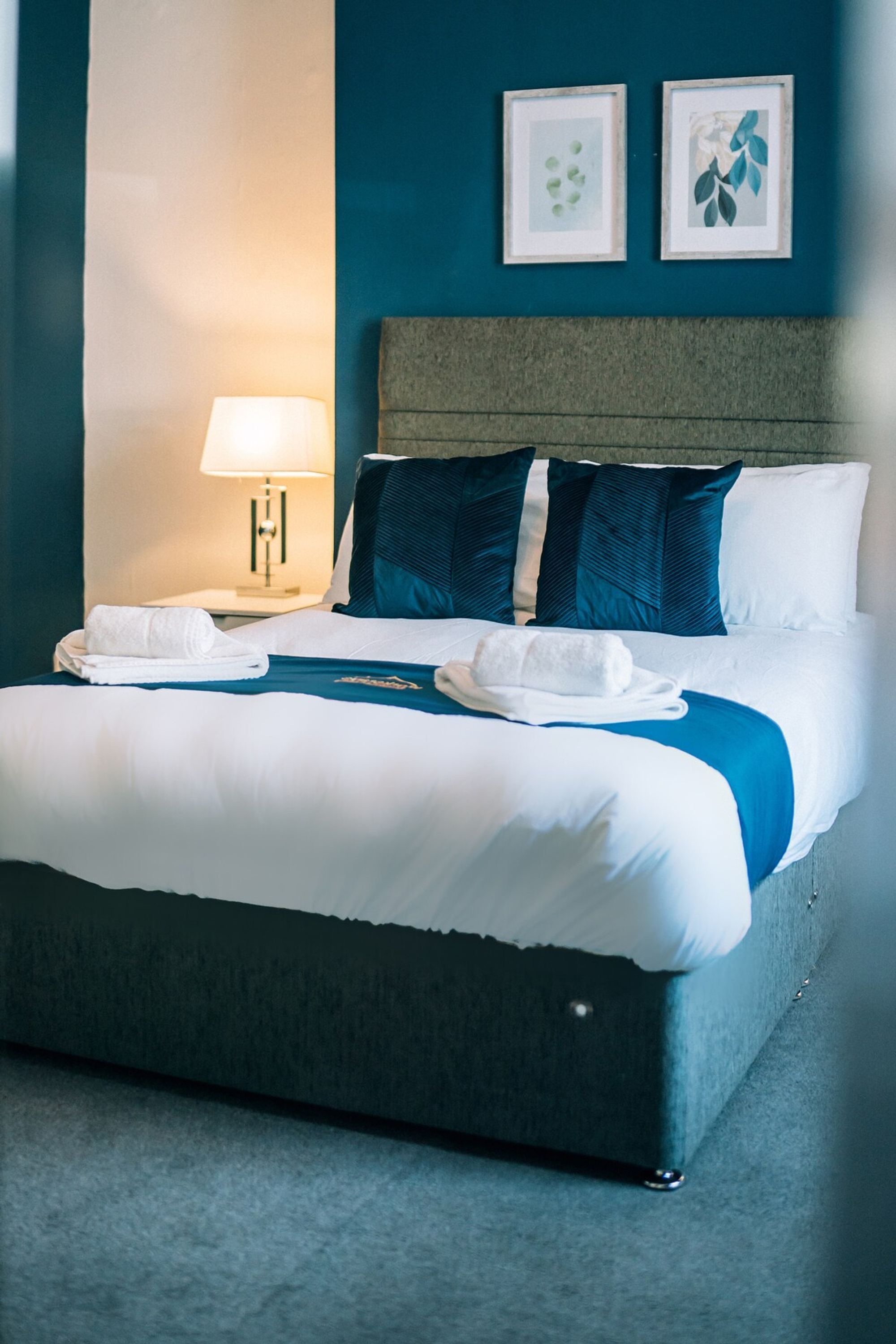 Une chambre d’hôtel avec un grand lit, une table de chevet avec une lampe et deux tableaux encadrés au mur.