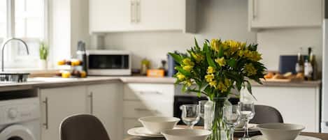 Una cocina moderna con armarios blancos, una mesa de comedor con cuencos y vasos blancos, y un florero con flores amarillas.