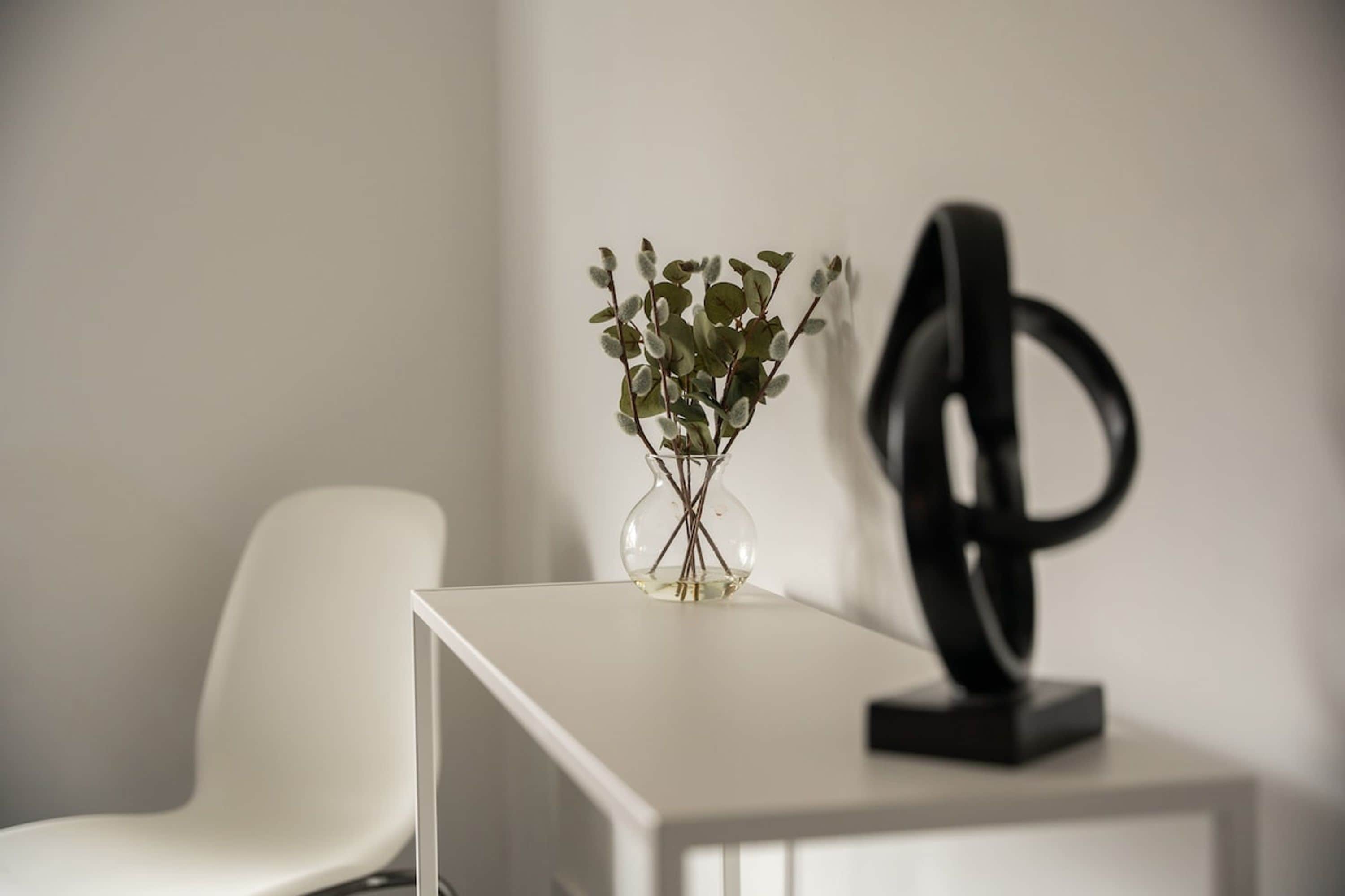 A white table with a clear glass vase containing green leaves and a black abstract sculpture.