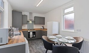 A compact kitchen with grey cabinets, a stainless steel range hood, and a built-in oven. There is a dining area with a glass table and four chairs. - Lord House by Horizon Stays (Redcar)