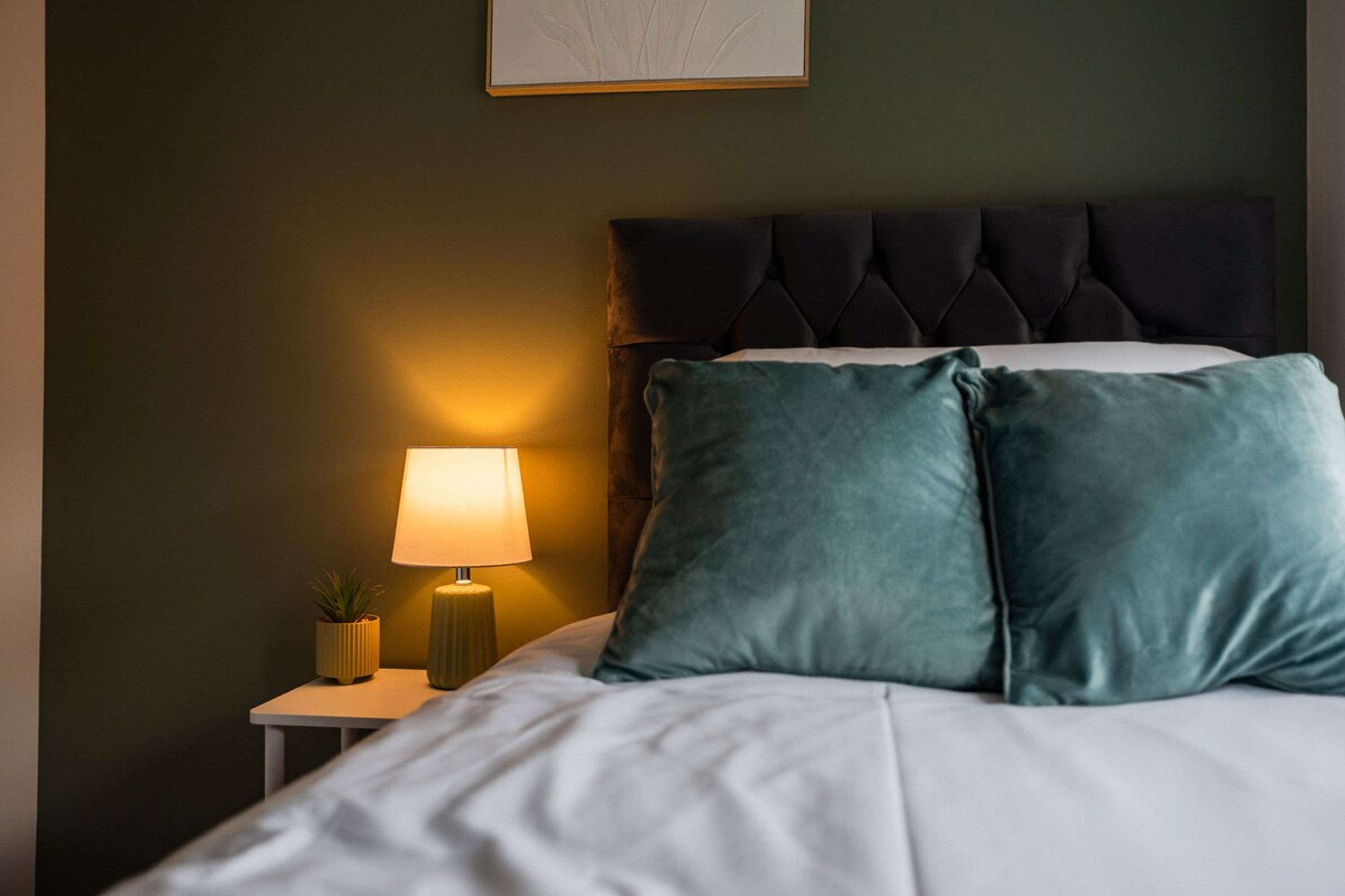 A neatly made bed with two pillows, a bedside lamp, and a small potted plant on a nightstand.
