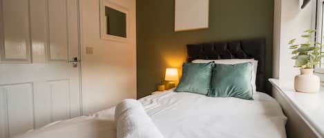 A neatly made bed with a white duvet and a rolled white towel, a dark headboard, a bedside lamp, and a potted plant on a windowsill.