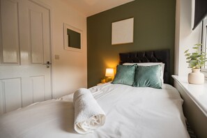 A neatly made bed with a white duvet and a rolled white towel, a dark headboard, a bedside lamp, and a potted plant on a windowsill. - Einstein House Horizon Stays (Stockton-on-Tees)