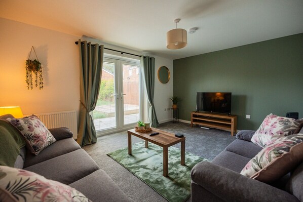 A living room with a sofa set, a coffee table, a television on a wooden stand, and a sliding glass door leading to an outdoor area. - Einstein House Horizon Stays (Stockton-on-Tees)