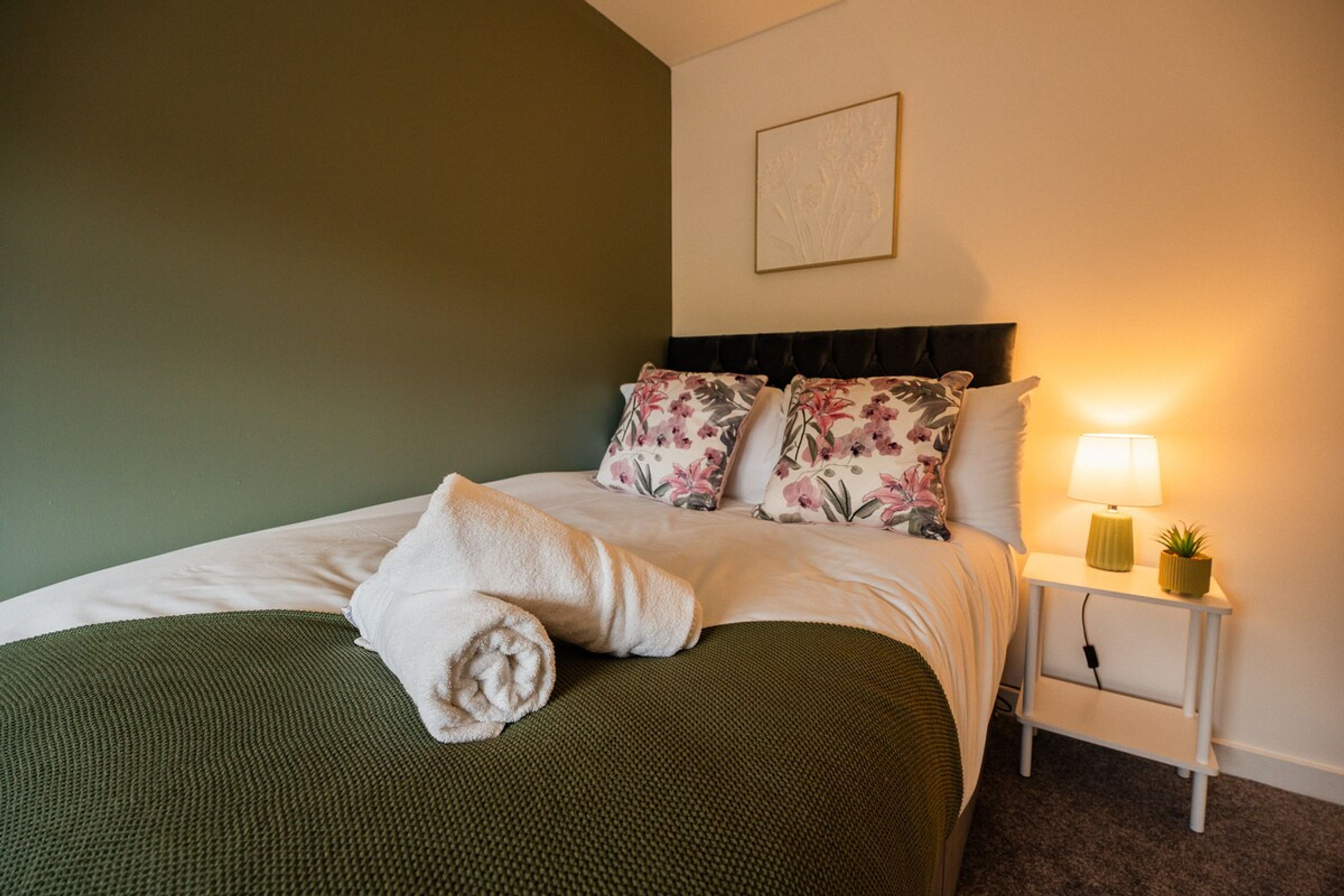 A neatly made bed with a rolled towel, a floral pillow, and a green throw blanket.