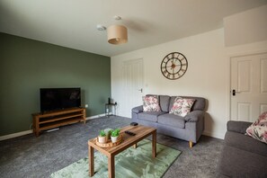 Ein Wohnzimmer mit einer Couch, einem Couchtisch mit Pflanzen, einem Fernseher und einer Wanduhr.
