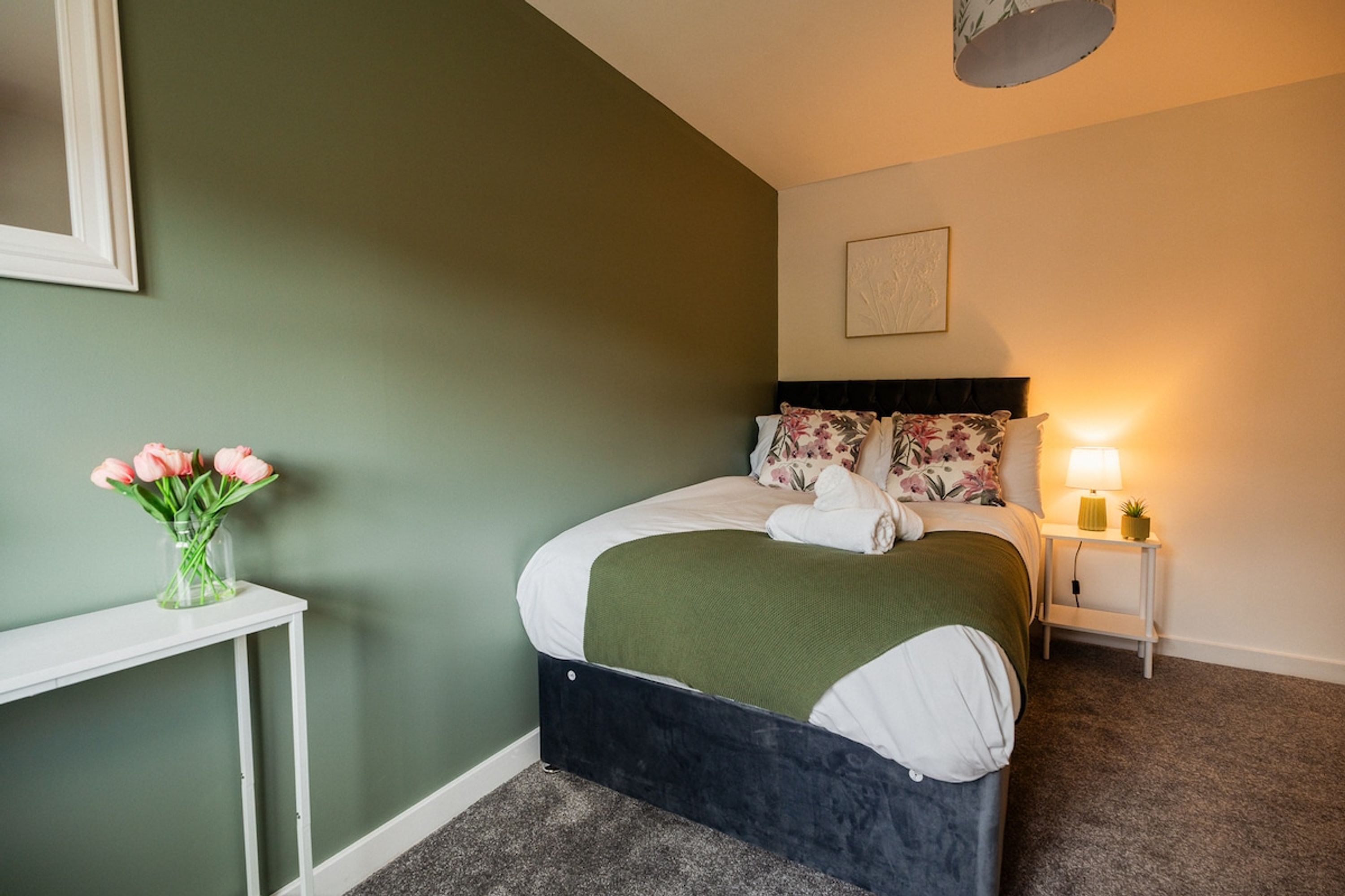 A bedroom with a bed, a bedside table, a nightstand, a lamp, and a vase of flowers.