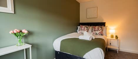 A bedroom with a bed, a bedside table, a nightstand, a lamp, and a vase of flowers.