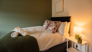 Una cama bien tendida con almohadones florales, una lámpara de noche y una pequeña planta en maceta sobre una mesita blanca.