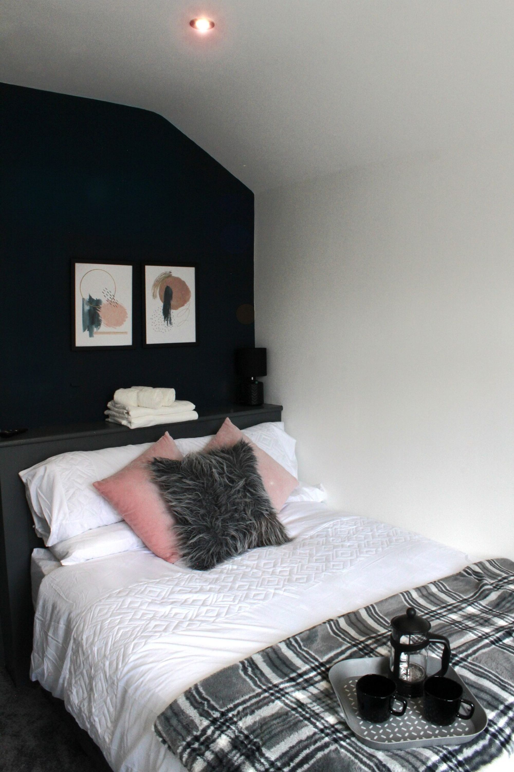 A bedroom with a bed, pillows, a blanket, a bedside table with a lamp, and artwork on the wall.