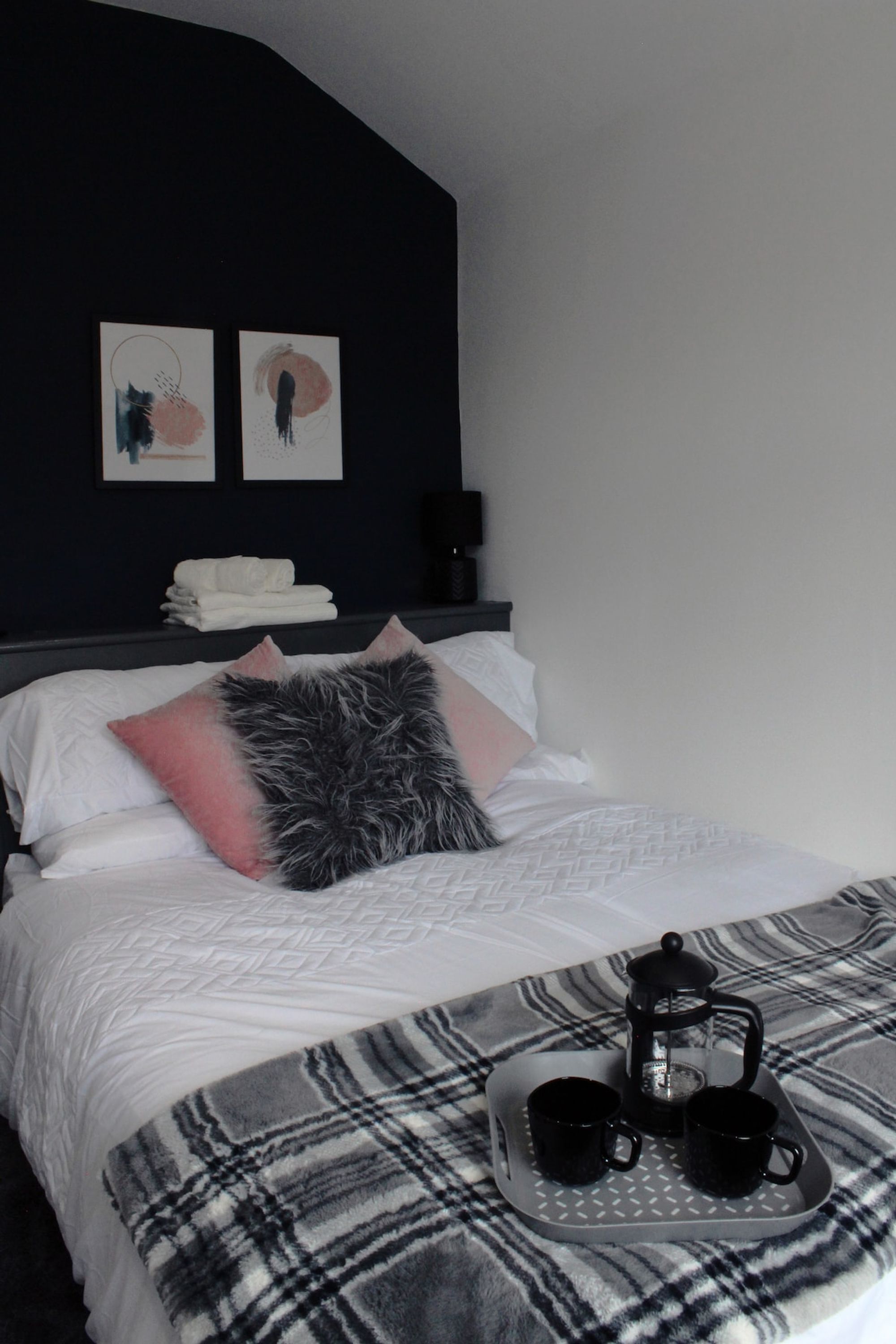 A hotel room with a bed, pillows, a tray with cups and a teapot, and artwork on the wall.