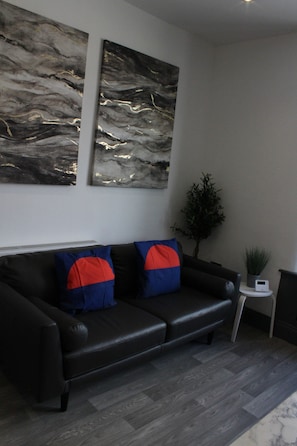 Ein modernes Wohnzimmer mit einem schwarzen Ledersofa, zwei abstrakten Gemälden, einem kleinen Beistelltisch mit einer Pflanze und einer digitalen Uhr.