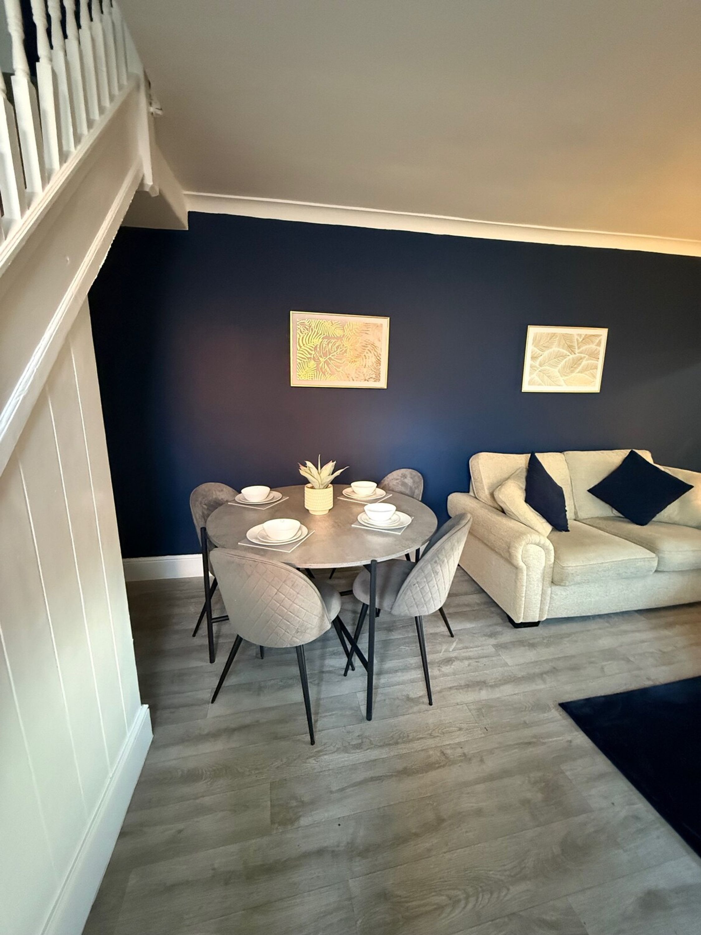 A dining area with a round table, chairs, and a white vase. There is a sofa with cushions in the background.