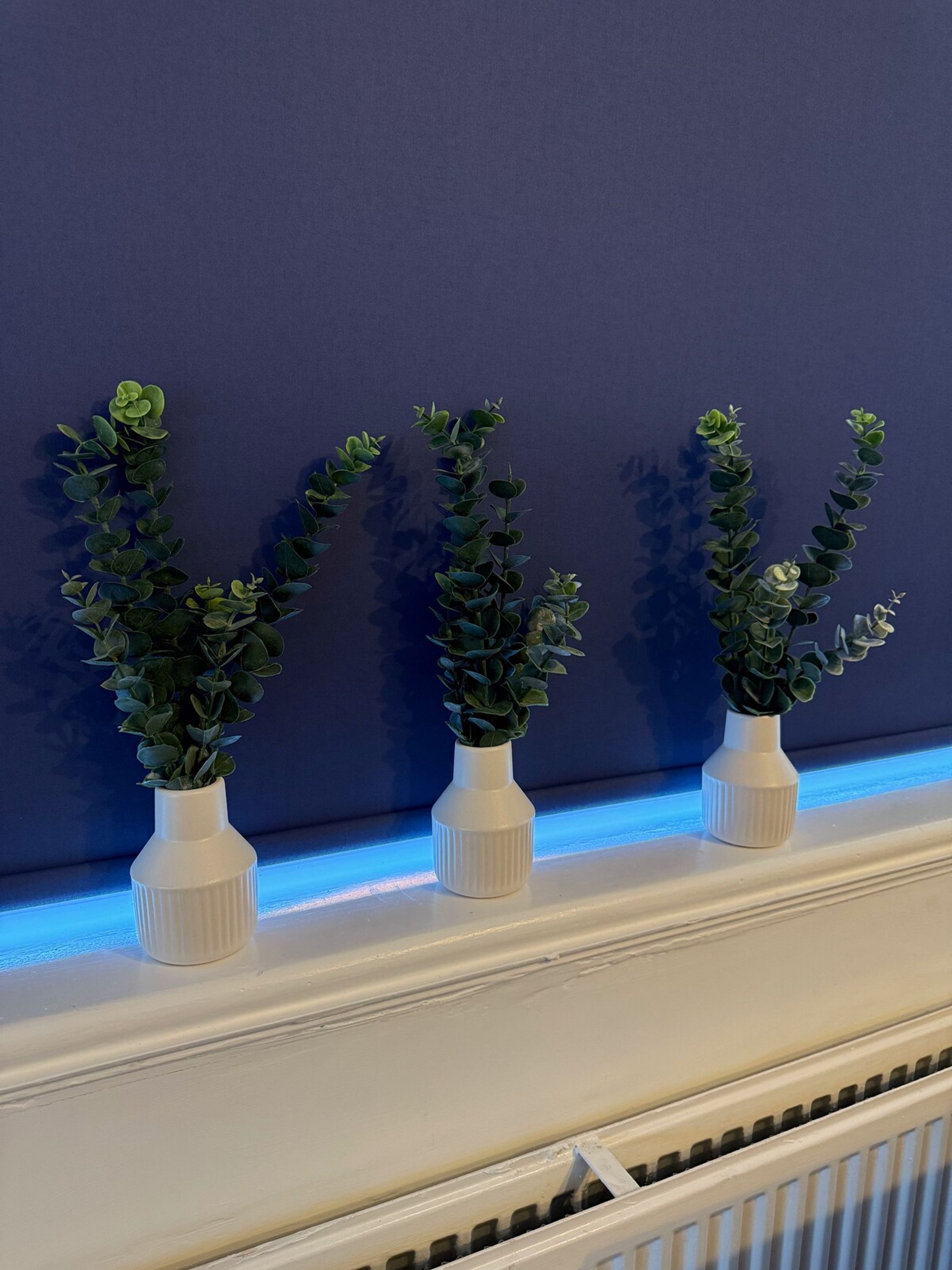Drie witte vazen met groene planten op een witte schoorsteenmantel tegen een donkere muur.