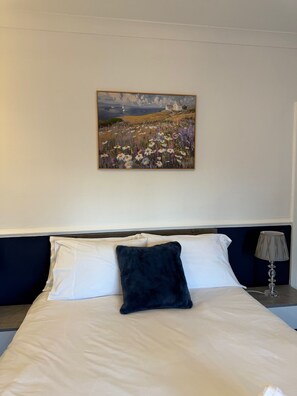 Habitació d'hotel amb un llit, un almoiner blau fosc, una tauleta de nit amb una làmpada i un quadre de paisatge a la paret.