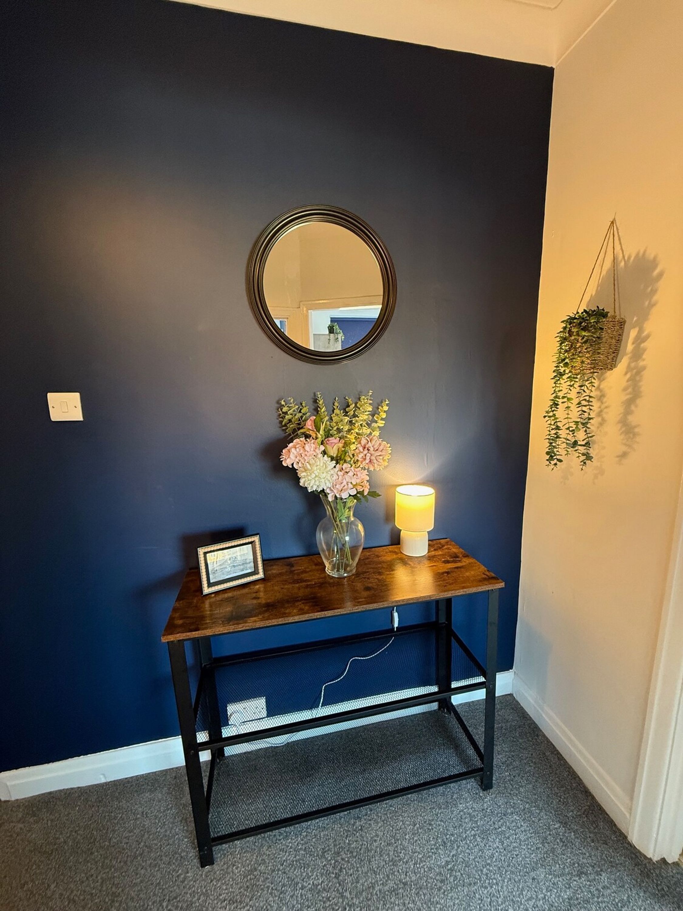 Una pared azul oscuro con un espejo redondo, un florero con flores, un cuadro enmarcado y una vela encendida sobre una consola de madera.
