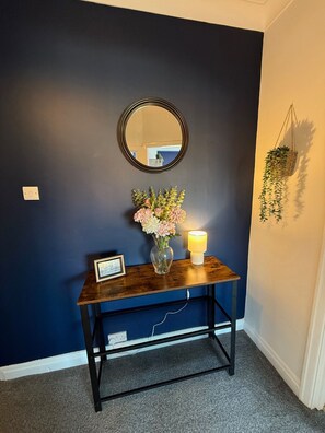 A dark blue wall with a round mirror, a vase with flowers, a framed picture, and a lit candle on a wooden console table. - Rochester House Horizon Stays (Middlesbrough)