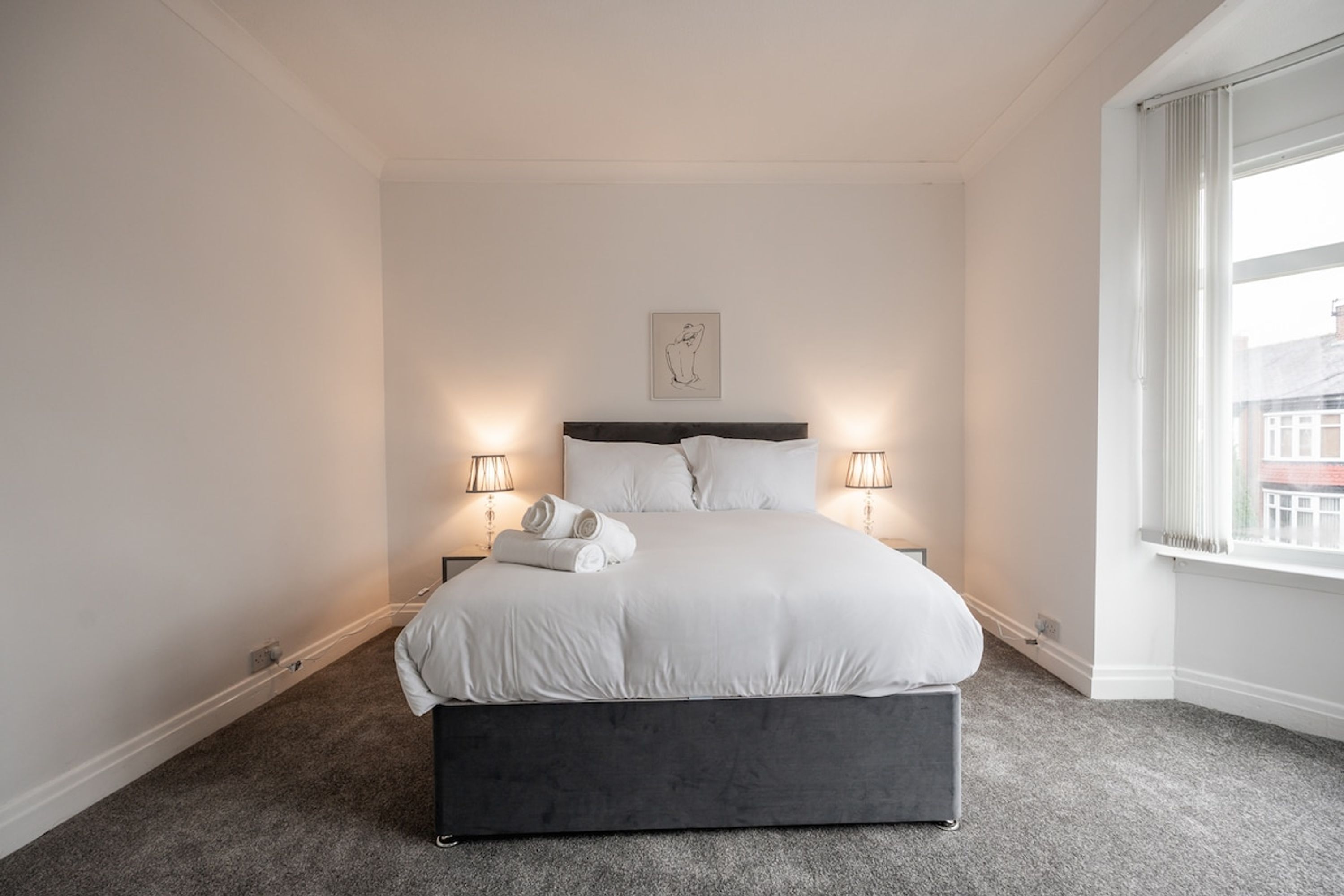 A bedroom with a bed, two bedside lamps, a window, and a wall-mounted artwork.