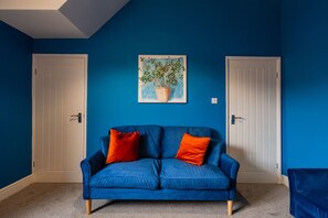 A room with a blue wall, a blue sofa with two orange cushions, and a painting of a plant on the wall. - The Blue Ember By Horizon Stays (Middlesbrough)