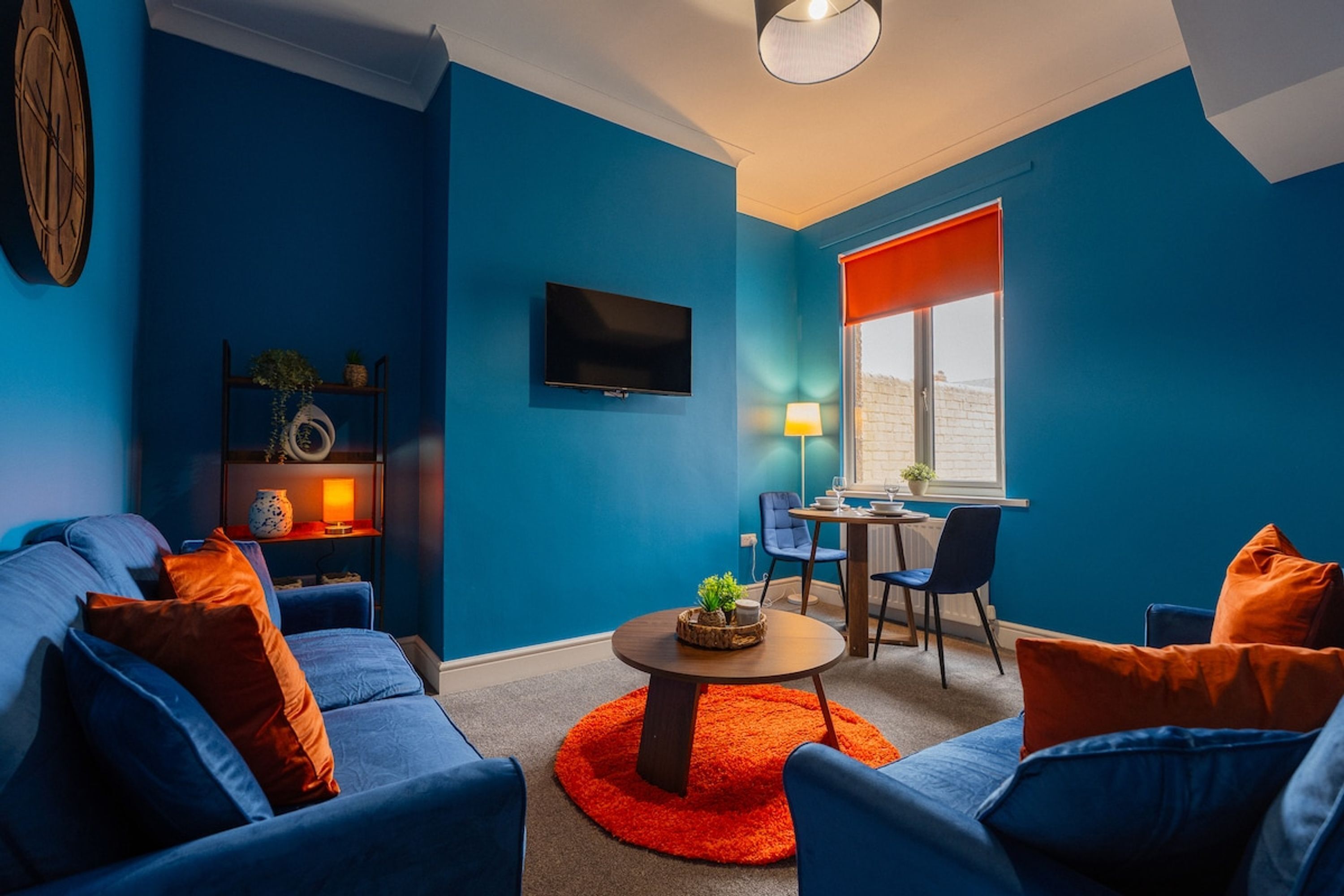 A modern living room with a blue wall, a flat-screen TV, a dining area with a table and chairs, and a cozy seating arrangement with sofas and a coffee table.