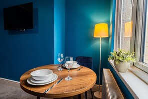 A dining area with a round table set for two, a flat-screen TV mounted on the wall, a floor lamp, and a window with a view. - The Blue Ember By Horizon Stays (Middlesbrough)