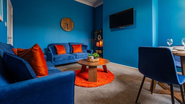Ein modernes Wohnzimmer mit blauer Wand, einem runden Holztisch, einem blauen Sofa mit orangenen Kissen, einem Essbereich mit Tisch und Stühlen sowie einem an der Wand befestigten Flachbildfernseher.