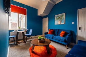 A room with a blue wall, a blue sofa with orange cushions, a wooden coffee table with a plant, a dining table with chairs, and a window with a red blind. - The Blue Ember By Horizon Stays (Middlesbrough)