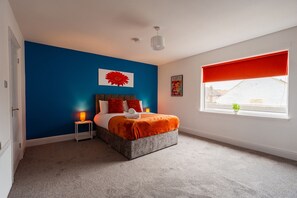 A bedroom with a bed, a nightstand, a window with a red blind, and a wall with a red flower decoration. - The Blue Ember By Horizon Stays (Middlesbrough)