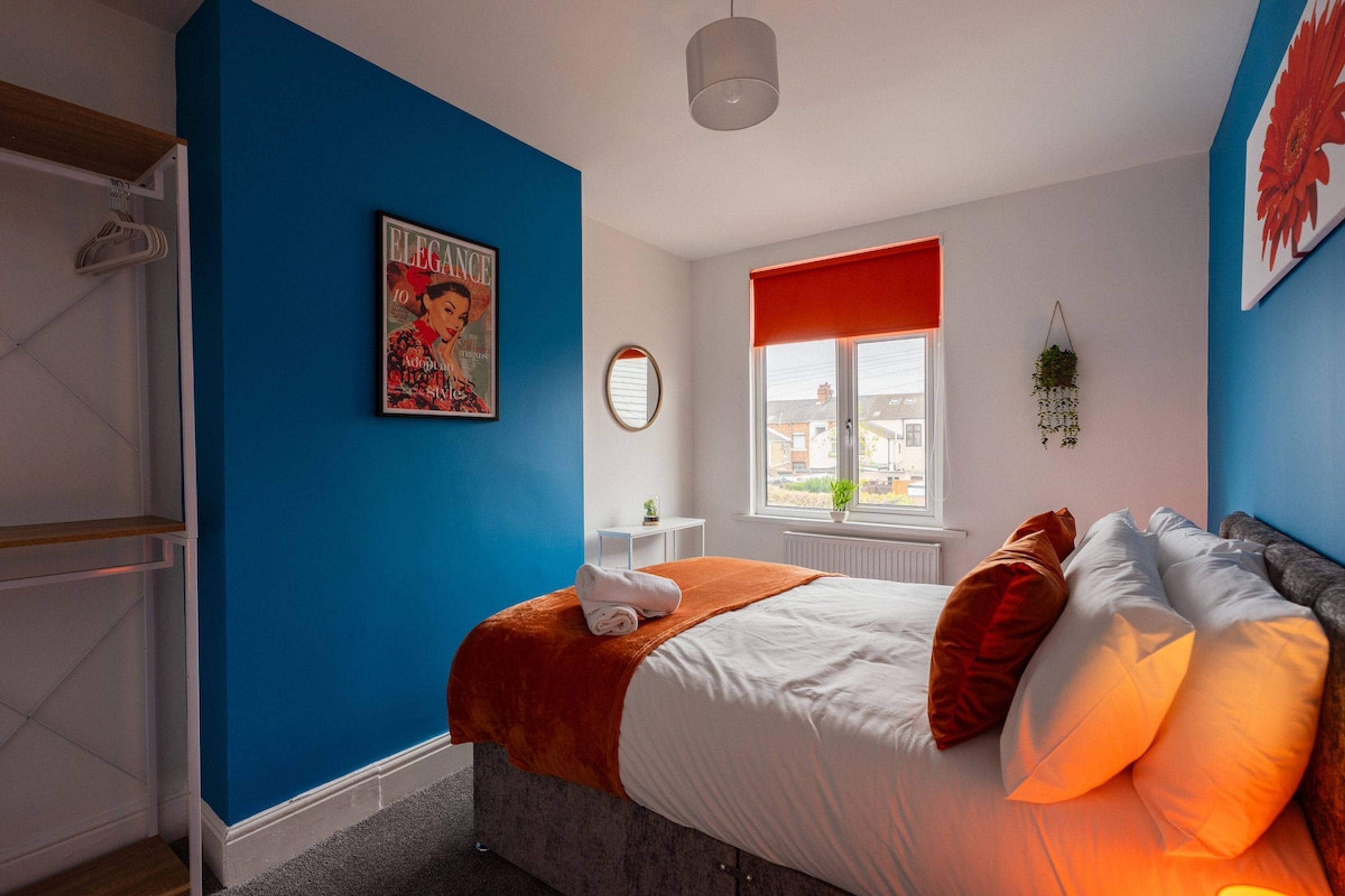 Une chambre à coucher avec un lit, une fenêtre avec un store rouge, un miroir fixé au mur et une affiche.
