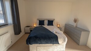 Ein Schlafzimmer mit Bett, Nachttischen, einem Schminktisch, einem Heizkörper und einem Fenster mit Vorhängen.