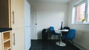 A compact kitchen with a dining area, featuring a white door, a small trash bin, a table with two chairs, and a window with a plant. - Thornwood House Horizon Stays (Stockton-on-Tees)