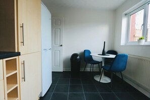 Eine kompakte Küche mit Essbereich, mit einer weißen Tür, einer kleinen Mülltonne, einem Tisch mit zwei Stühlen und einem Fenster mit Pflanze.