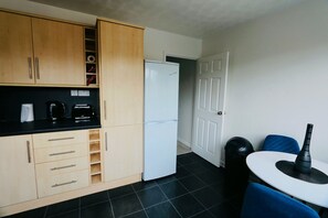 A compact kitchen with wooden cabinets, a white refrigerator, a black coffee maker, and a dining table with a blue chair. - Thornwood House Horizon Stays (Stockton-on-Tees)