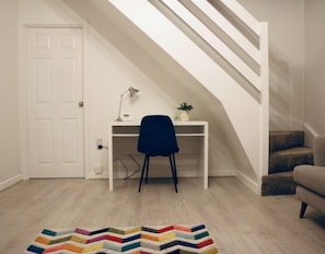 Ein Zimmer mit weißer Tür, einem Schreibtisch mit Stuhl, einer kleinen Pflanze und einer Treppe.