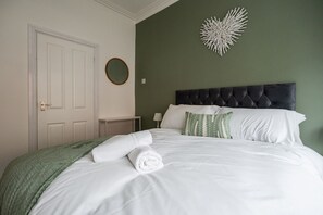 A neatly made bed with white linens and a green throw blanket, a round mirror on the wall, and a tufted headboard. - The Alfred Suite Redcar (Redcar)
