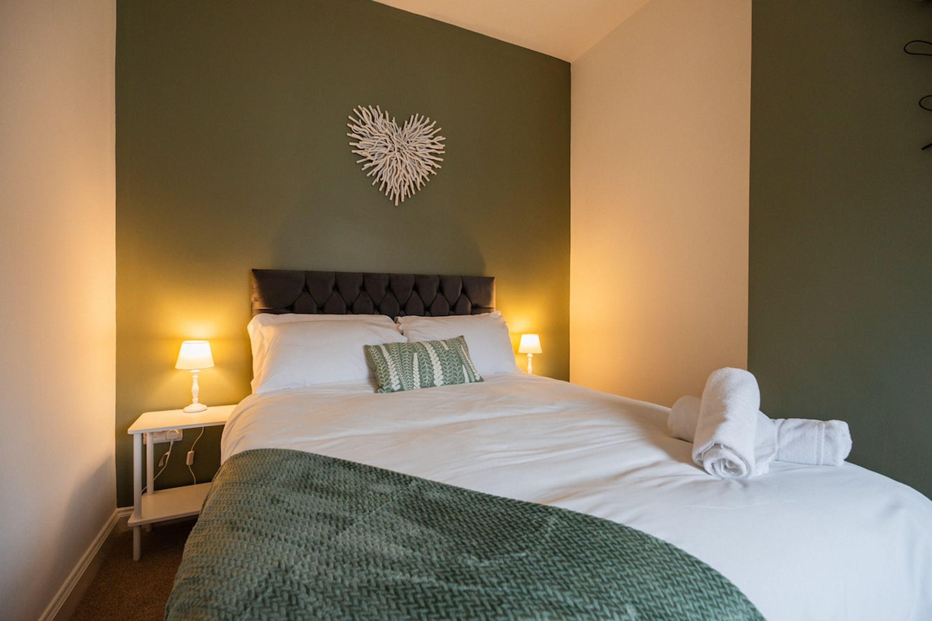 Een net opgemaakt bed met witte lakens en een groene plaids, twee bedlampen en een decoratief kunstwerk erboven.
