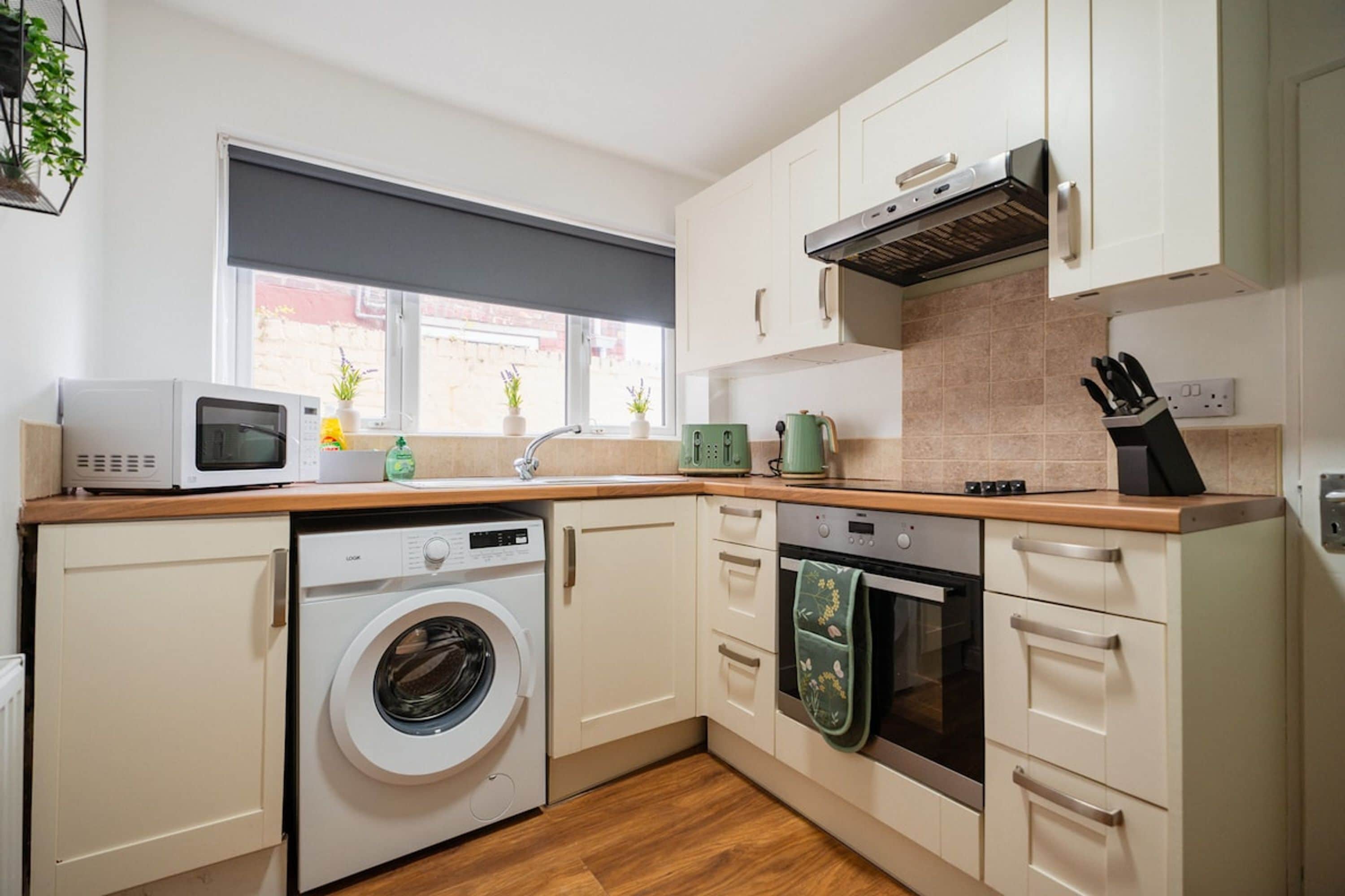 A compact kitchen with a washing machine, microwave, oven, and a window with a view of the street.