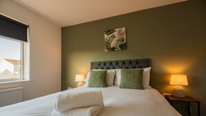 Ein ordentlich bezogenes Bett mit weißen Leinen, zwei grünen Kissen und einem gefalteten weißen Handtuch. Auf den Holz-Nachttischen stehen je eine Nachttischlampe.