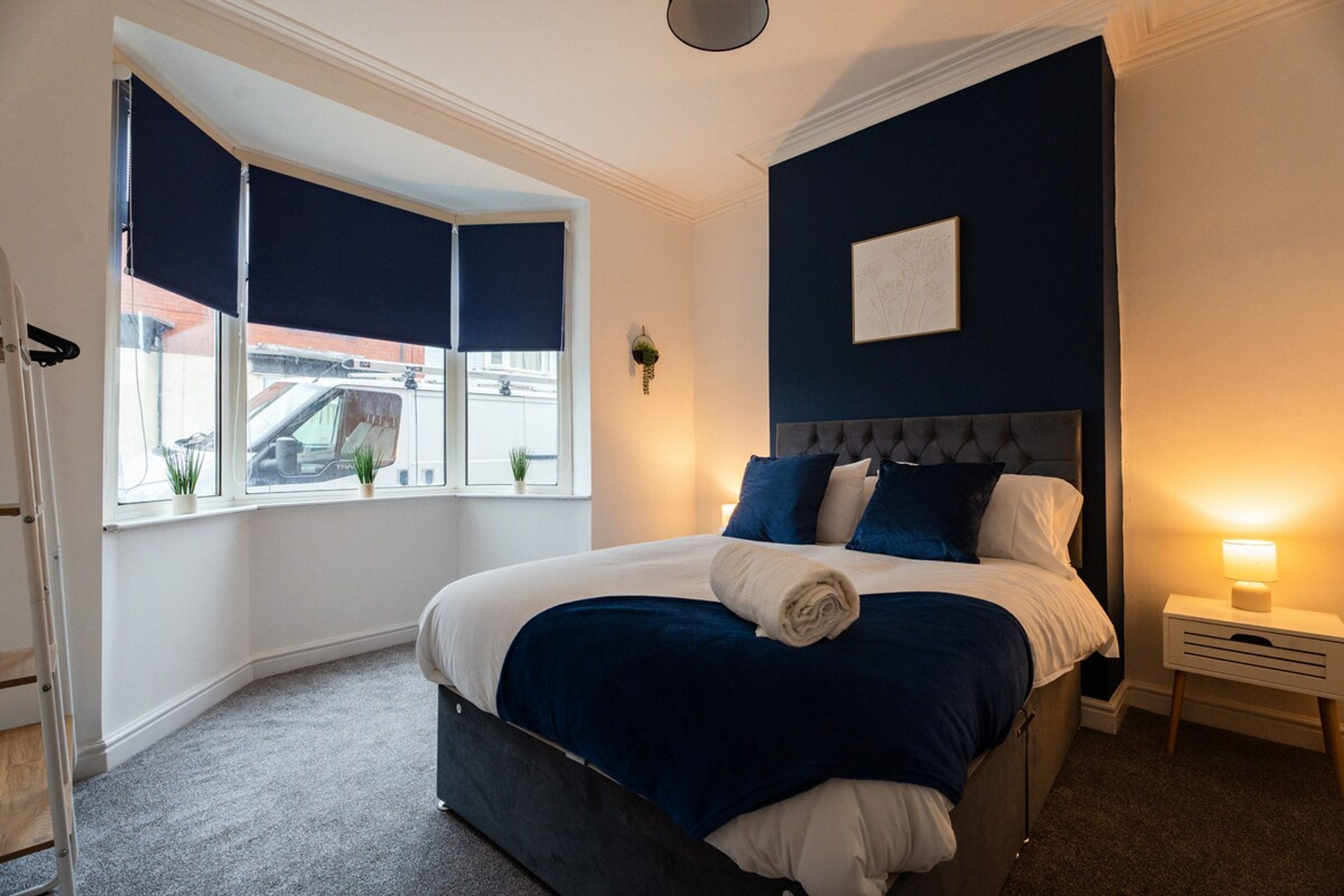 Ein Schlafzimmer mit Bett, Nachttisch, einem Fenster mit Jalousien und einem Wandbild.