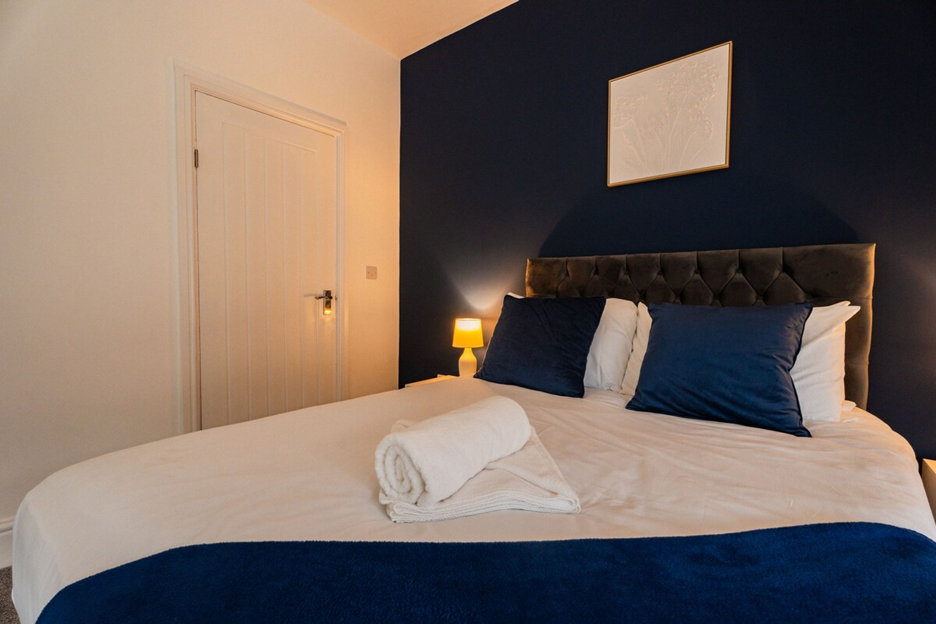 A hotel room with a bed, a door, a nightstand with a lamp, and a framed picture on the wall.