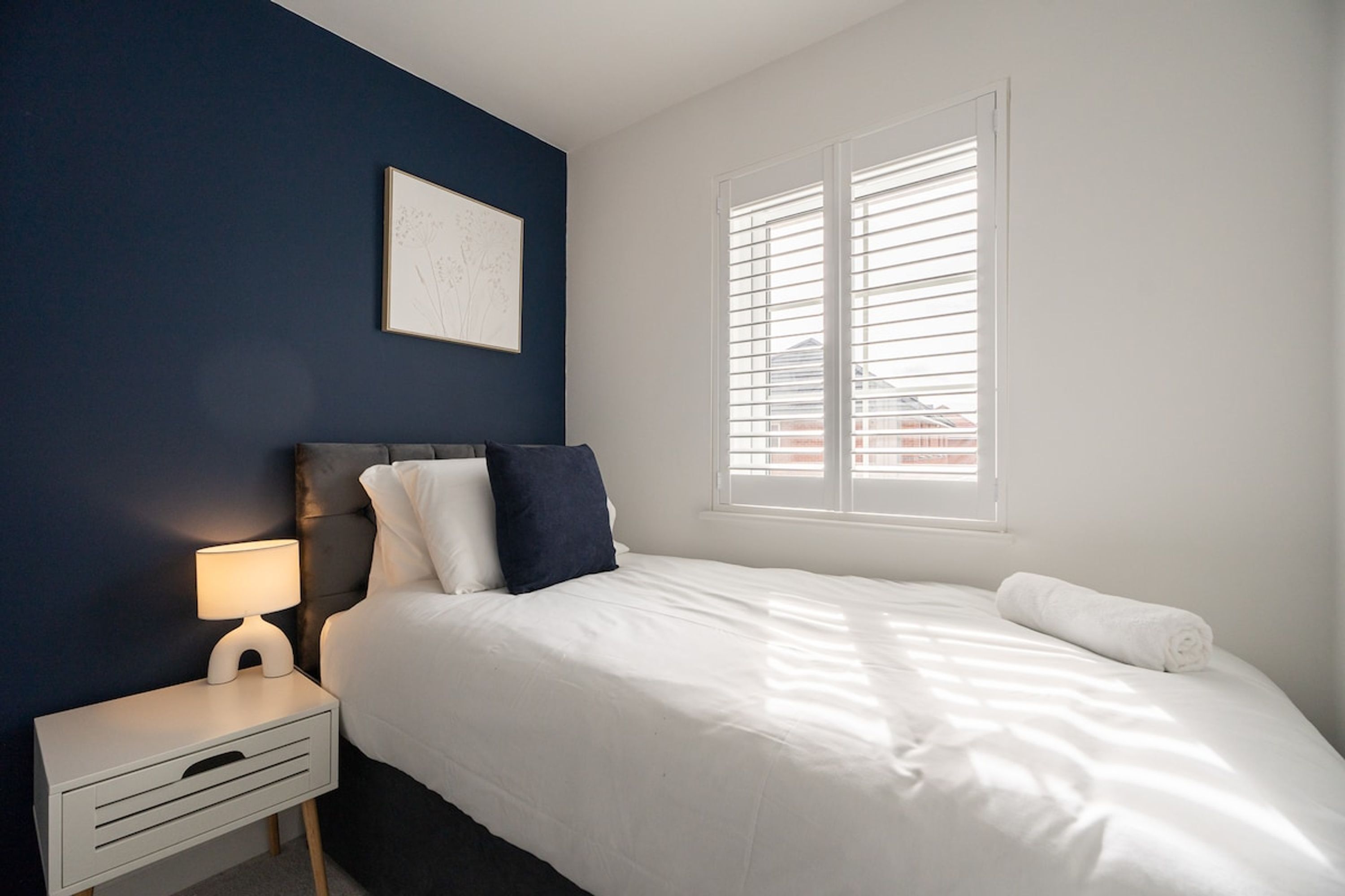 Een slaapkamer met een bed, nachtkastje, lamp, raam met jaloezieën en een lijst aan de muur.